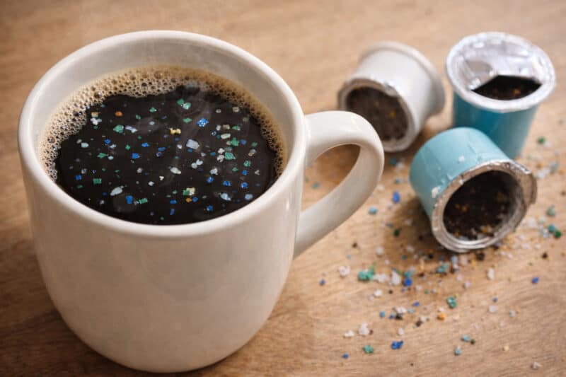 Cup of coffee with visible microplastic particles on the surface beside used plastic K-cup pods, illustrating how plastics can enter coffee during brewing.