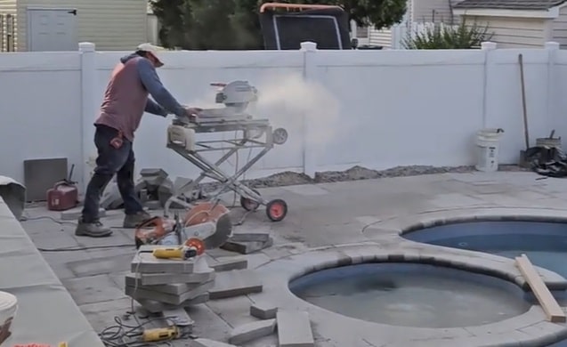 Worker using a masonry saw cutting concrete pavers with visible silica dust cloud spreading in a residential backyard near a pool.