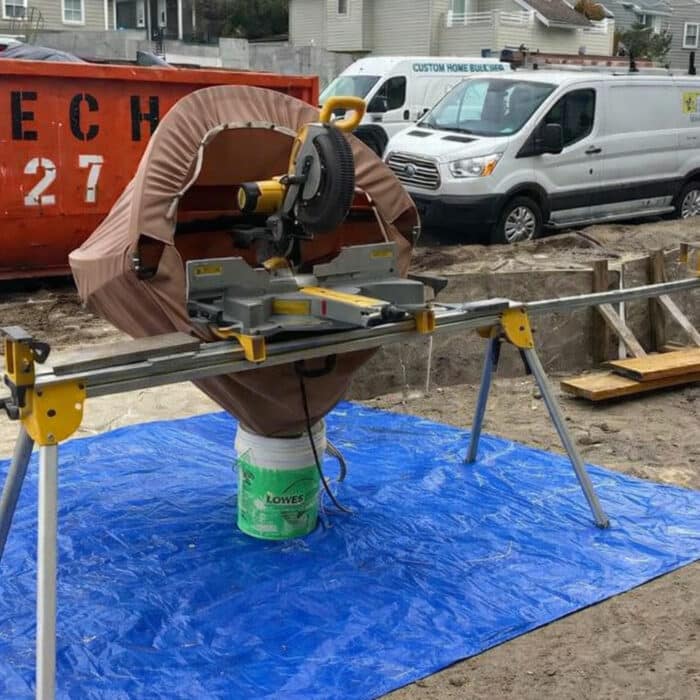 Miter saw with dust containment hood positioned over a blue tarp on a residential construction site to capture debris and prevent dust pollution.