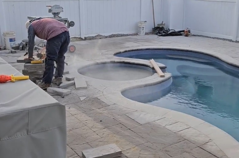A medium shot of a male construction worker, wearing a mask and safety glasses, cutting concrete pavers with an angle grinder on a residential pool deck. He is bending over, and a large cloud of fine white dust is pluming upwards. Behind him is an empty swimming pool shell with a winding design and a hot tub. In the far background, beyond a white fence, are other houses under a clear sky. Scattered tools and a table saw on a stand are visible on the dusty patio.